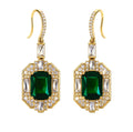 A pair of gold drop earrings featuring large emerald-cut green center stones surrounded by clear baguette and round crystals, set against a plain white background.
